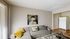 Modern living room with a stylish gray sofa adorned with vibrant yellow and patterned cushions, contemporary art on the wall, and an inviting atmosphere near the kitchen area with natural light streaming through vertical blinds.