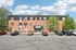 Charming brick apartment building with sleek black roofing, surrounded by lush greenery and ample parking space under a vibrant blue sky.