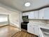 Bright kitchen with modern white cabinets, marble countertops, and stainless steel appliances opening into a sunlit living area with wooden floors.