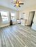 Bright and airy room with wooden floors, large windows, and a modern ceiling fan, showcasing an inviting open-concept layout with kitchen access.