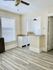 Bright open area featuring large windows with blinds, stylish ceiling fan, and modern wood flooring. Adjacent to a compact kitchen with white cabinetry and red pendant lighting, perfect for intimate home living.