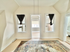 Bright, inviting room with large windows and elegant drapes complemented by a decorative rug on light flooring, leading to a sunlit nook through a charming doorway.