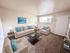 Bright and inviting living room with elegant beige sofas, soft blue accent pillows, and a modern coffee table. Natural light pours through large windows, enhancing the cozy, carpeted space.