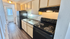 Modern kitchen with sleek black appliances, ample countertop space, and wood cabinetry, leading to a well-lit hallway with bathroom access.