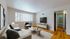 Bright and inviting living room with modern decor, featuring a comfortable sofa, stylish coffee tables, and a large flat-screen TV; natural light streaming through a window with vertical blinds enhances the warm wood flooring.