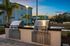 Elegant outdoor grilling area with modern stainless steel grills and lush landscaping at sunset.