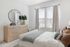 Bright bedroom with a light wood dresser, round mirror, and comfortable bed with cozy pillows and blanket.