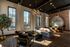 Elegant living area with modern decor, featuring large windows, stylish chandelier, and comfortable seating, adjacent to a brick-accented workspace under high ceilings.