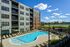 Luxurious apartment complex featuring a sparkling outdoor pool surrounded by elegant lounge chairs and vibrant greenery, offering scenic river and city views under a bright blue sky.