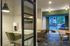 Contemporary apartment interior featuring a cozy study nook with a green chair and modern lighting, leading to a stylish dining area with elegant seating and large windows offering vibrant city views.