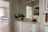 Modern bathroom with sleek white cabinetry and a large mirror, adjacent to a stylish kitchen area with elegant pendant lighting and subtle decor accents.