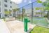 Modern apartment complex with a tennis court, barbecue area, and lush landscaping under a clear blue sky.