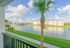 Bright apartment balcony view featuring a serene lake, manicured lawns, and modern residential buildings under a clear blue sky.