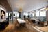 Luxurious open-concept apartment with floor-to-ceiling windows offering stunning city views, featuring a stylish dining area with a long marble table and modern chairs, spacious living room with contemporary furnishings, and elegant wooden flooring.