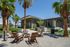 Modern outdoor communal area with palm trees, cozy seating around a fire pit, and a sleek barbecue grill next to a contemporary building.
