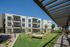 Modern apartment building with balconies, set in a landscaped garden featuring hammocks and lush palm trees under a clear blue sky.