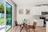 Bright dining area with modern glass table, elegant brown chairs, and a sleek kitchen featuring stainless steel appliances and white cabinetry, opening to a lush green outdoor view.