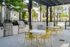Modern outdoor patio area featuring sleek stainless steel grills, vibrant yellow chairs, and a scenic view of a landscaped poolside with palm trees.