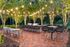 Charming outdoor patio with string lights, modern seating, and stylish grills set on a vibrant brick floor, surrounded by lush greenery.