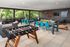 Modern outdoor entertainment space with sleek foosball table, turquoise chairs, cozy seating area, and vibrant greenery overlooking lush trees.