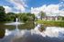 Charming apartment complex overlooking a serene pond with a stunning fountain, surrounded by lush greenery and blue skies.