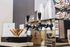 Modern beverage dispenser with sleek black taps, surrounded by neatly stacked clear plastic cups and a holder with stirring sticks, offering a chic refreshment station.