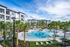 Luxurious apartment complex with a stunning resort-style pool, surrounded by lush palm trees and modern buildings under a clear blue sky.
