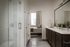 Elegant bathroom with wood cabinetry, modern fixtures, and glass-enclosed shower leading to a cozy, sunlit bedroom.
