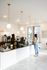 Modern cafe interior with sleek white tiled counter, elegant pendant lighting, and a welcoming barista serving a customer in casual attire.