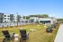 Outdoor amenity area at an apartment community featuring a large lawn with cornhole boards and Adirondack chairs, overlooking a fenced swimming pool and clubhouse.