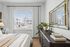 Bright and elegant bedroom with natural light, featuring a cozy bed, stylish dresser with books and decor, and a large mirror reflecting a modern apartment view.
