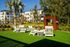Charming outdoor lounge area with modern white chairs on lush green grass, surrounded by tropical palm trees and contemporary apartment buildings.