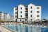 Luxury apartment complex featuring a sunlit swimming pool with sun loungers and umbrellas, surrounded by modern multi-story buildings.