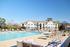 Luxurious apartment complex with a sparkling pool, sun loungers, and shaded seating areas under a clear blue sky.