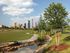 Scenic view of a lush green park with a pond and city skyline in the background.