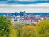Panoramic view of a bustling city skyline with lush greenery in the foreground and a dramatic cloudy sky.