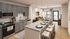 Modern kitchen with sleek gray cabinets and granite countertops, leading into a cozy, well-lit living room with stylish decor.