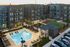 Modern apartment complex with a large swimming pool, surrounded by lounge chairs and lush landscaping. The buildings feature multiple stories with private balconies overlooking the courtyard.