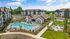 Luxury apartment complex with a large sparkling swimming pool, surrounded by lush gardens and modern buildings under a bright blue sky.