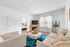 Bright and inviting living room with modern beige sofas, a vibrant blue rug, and a wooden fireplace, leading to a cozy bedroom.