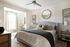 Elegant bedroom featuring a plush bed with stylish cushions, a sunlit window nook with light drapes, and sophisticated decor including a round mirror and framed art.