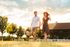 Happy couple strolling through a sunlit garden, holding hands and enjoying a picturesque day with a rustic house in the background.