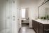 Luxurious modern bathroom with sleek glass shower, elegant vanity with double sinks, and view into a bright, cozy bedroom.