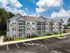 Contemporary three-story apartment building with elegant balconies, set against a bright blue sky, surrounded by lush greenery and ample parking space.