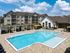 Luxurious apartment complex featuring a sparkling pool with surrounding lounge area and modern architecture under a bright blue sky.