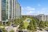 Modern high-rise apartment building overlooking a lush green park with a serene pond and walking paths, bathed in bright sunlight under a clear blue sky.