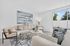 Bright and modern living room featuring elegant furnishings, large artwork, and a view of lush greenery through a floor-to-ceiling window.