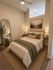 Cozy bedroom featuring a comfortable bed with elegant green accents, warm lighting, and a serene landscape painting above the headboard. Modern design with a full-length mirror and natural decor elements.