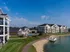 Elegant apartment complex with spacious balconies, lush green lawns, and a serene lakeside view under a clear blue sky.
