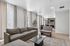 Modern open-plan living room with elegant gray sofas, chic glass coffee table, and expansive windows leading to a sleek kitchen and dining area.